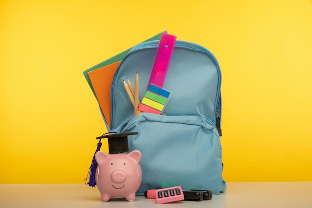 Bright backpack with school tools and piggy bank in hat. Savings for education concept