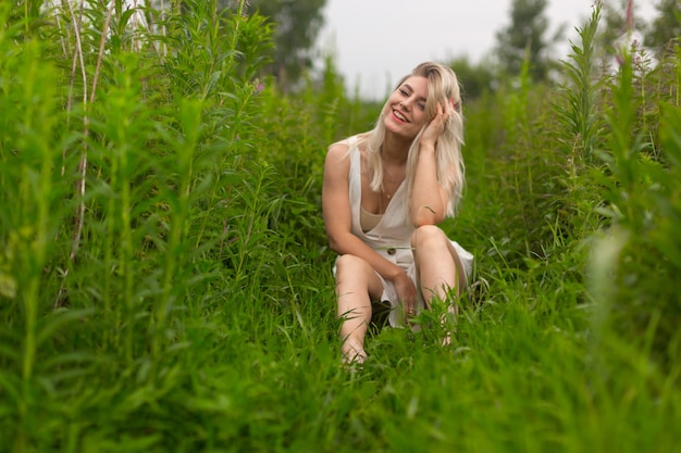 La ragazza bionda brillante in un vestito bianco estivo si siede tra l'erba alta.