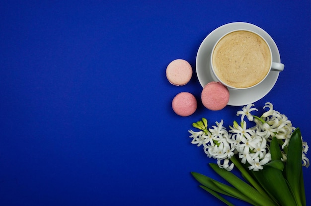 On a bright blue background there is a cup of cappuccino coffee white hyacinths and macaroni