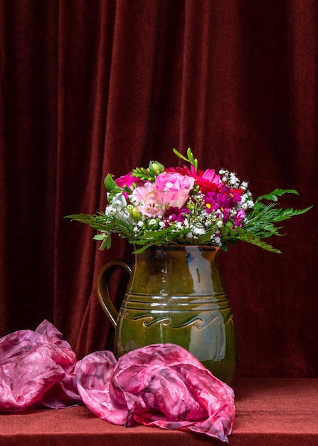 A bright bouquet of flowers in a green vase on a dark fabric background next to a pink scarf Still