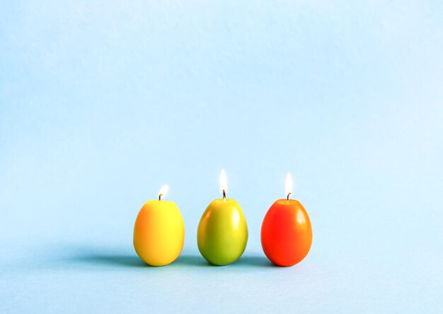 Bright burning paraffin candles in the shape of colorful Easter eggs on blue background.