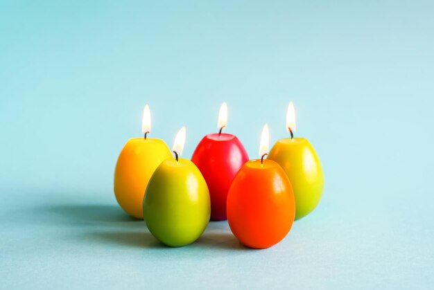 Bright burning paraffin candles in the shape of colorful Easter eggs on blue background.