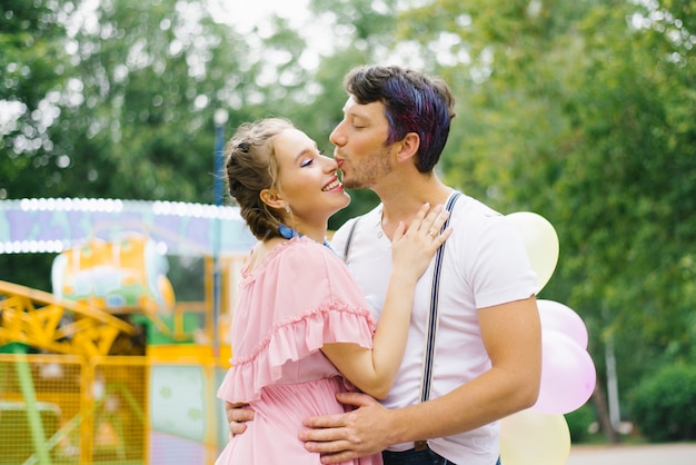 Coppie allegre luminose nell'amore che camminano di estate in un parco di divertimenti. il ragazzo bacia la sua ragazza sul naso, lei sorride di rimando