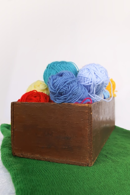 Bright colorful balls of yarn and knitteng needles in a wooden box on white background with green knitted mat.