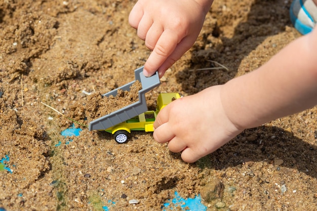 Bright colorful yellow green plastic toy truck in child hands, little kid is playing in sandbox with his truck toy