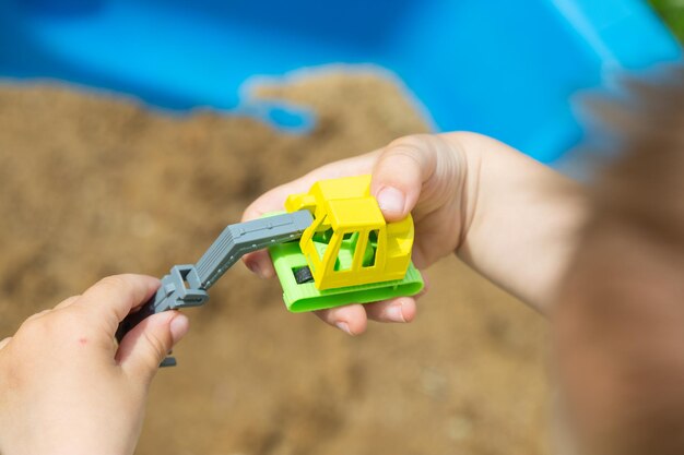 Bright colorful yellow green plastic toy truck in child hands, little kid is playing in sandbox with his truck toy