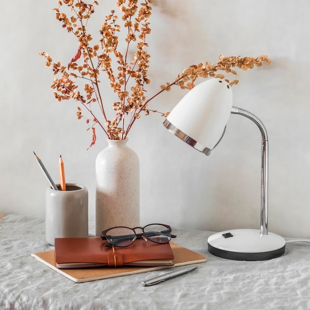 스칸디나비아 스타일의 밝고 아늑한 객실 세라믹 꽃병 연필에 테이블 램프 꽃이 있는 테이블 가죽 공책 및 안경