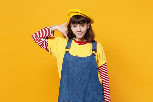 Bright girl teenager in french beret, denim sundress doing phone gesture like says call me back isolated on yellow background in studio. People sincere emotions, lifestyle concept. Mock up copy space.