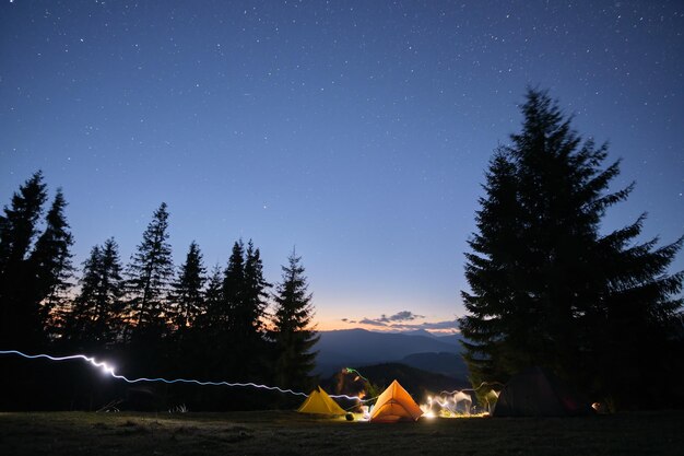 Bright illuminated tourist tents glowing on camping site in dark mountains under night sky with sparkling stars Active lifestyle concept