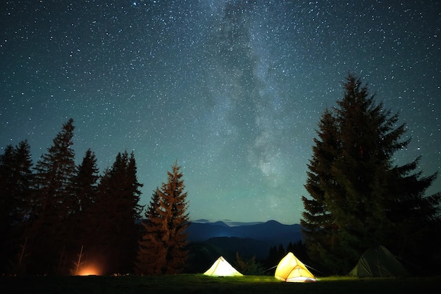 輝く星と夜空の下の暗い山の森のキャンプ場で、輝くたき火の近くに明るく照らされた観光テントアクティブなライフスタイルとアウトドアライフのコンセプト