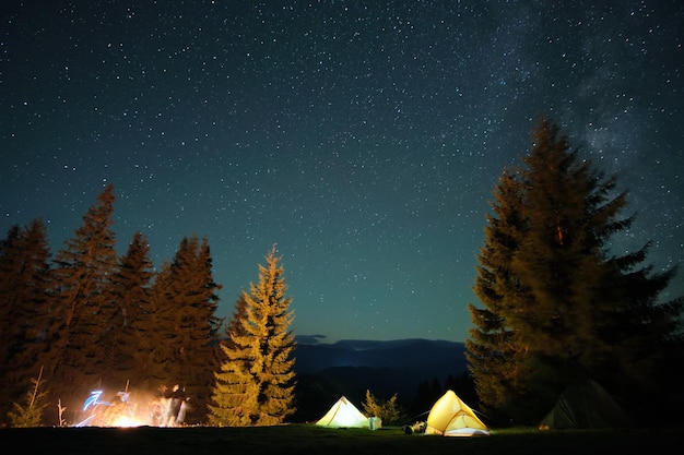 Bright illuminated tourist tents near glowing bonfire on camping site in dark mountain woods under night sky with sparkling stars Active lifestyle and outdoor living concept