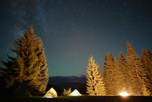 Bright illuminated tourist tents near glowing bonfire on camping site in dark mountains under night sky with sparkling stars Active lifestyle concept