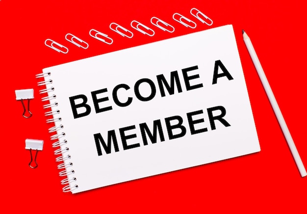 On a bright red background, a white pencil, white paper clips, and a white notebook with the text BECOME A MEMBER