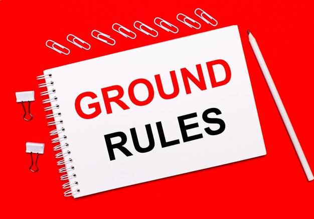 On a bright red background, a white pencil, white paper clips, and a white notebook with the text GROUND RULES.