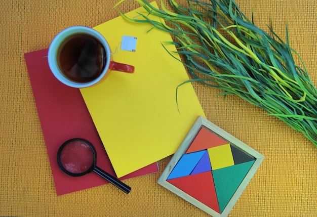 Lente d'ingrandimento della tazza del tè dei taccuini rossi e gialli luminosi e mockup dello spazio della copia del tangram