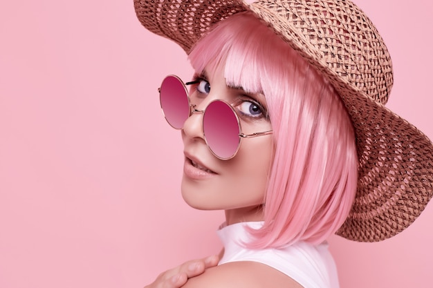 Foto ritratto estivo luminoso di una ragazza bellissima e positiva con capelli rosa, occhiali da sole e un cappello intrecciato in studio colorato