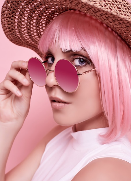 Foto ritratto estivo luminoso di una ragazza bellissima e positiva con capelli rosa, occhiali da sole e un cappello intrecciato in studio colorato