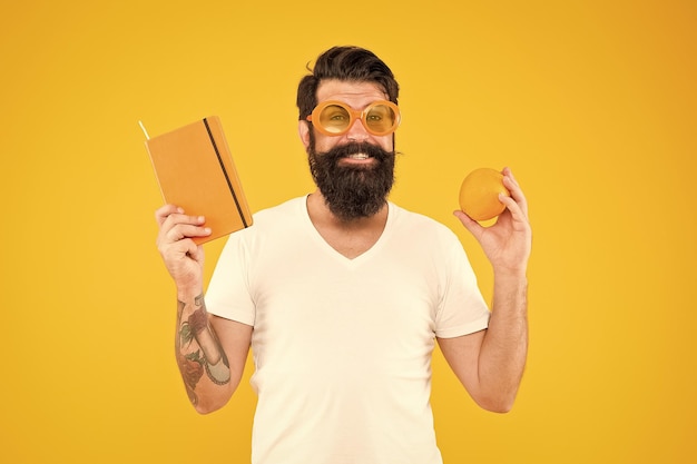 Bright and vibrant Man bearded cheerful smiling hipster wear orange color sunglasses while hold orange fruit and book notepad Total orange Guy long beard likes orange color Vitamin source