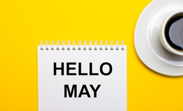 On a bright yellow background, a white cup with coffee and a white notepad with the words HELLO MAY