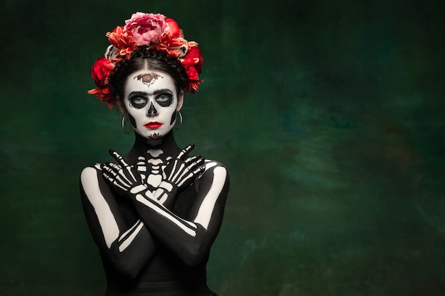 Bright. Young girl like Santa Muerte Saint death or Sugar skull with bright make-up. Portrait isolated on dark green studio background with copyspace. Celebrating Halloween or Day of the dead.