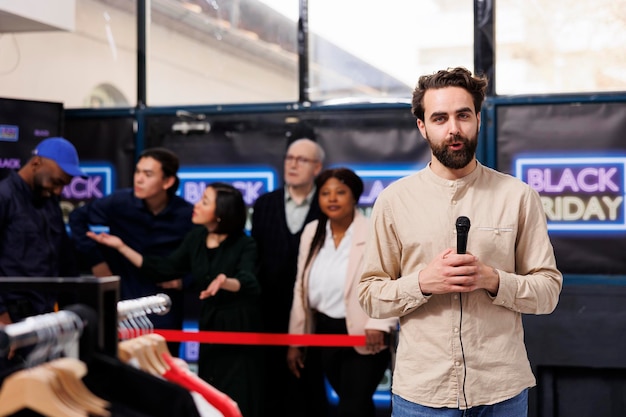 Broadcast journalist standing against crowd of eager shoppers waiting for Black Friday sales. TV reporter holding microphone telling at camera about shopping frenzy during seasonal sales