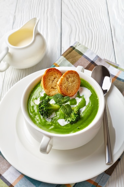 Broccoli cream soup with fresh mint in a white soup bowl served with toasted slices of baguette and fresh cream, vertical view from above