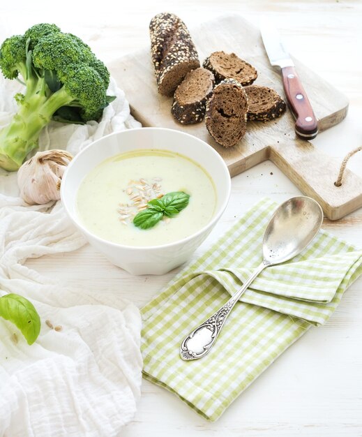 Broccoli crème soep met zonnebloempitten, verse basilicum en brood in kom over witte houten tafel