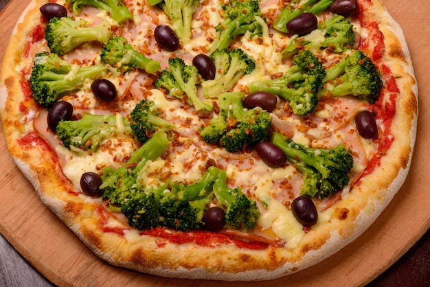 Broccolipizza met zwarte olijven op een houten bord en groenten op de achtergrond.