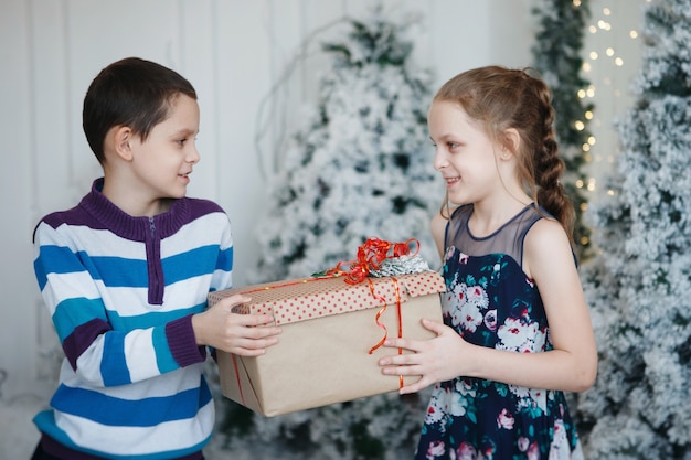 Broer en zus geven elkaar een geschenkdoos bij de kerstboom.