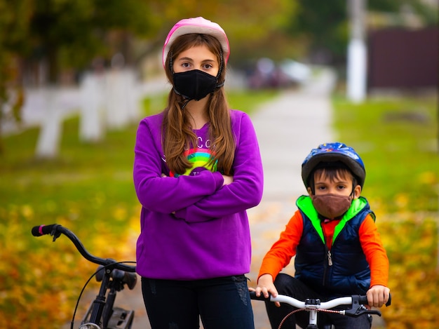 Broer en zus in het herfstpark, fietsen die beschermende maskers dragen. In de muur, een wazig herfstpark. Pandemie en virusconcept.