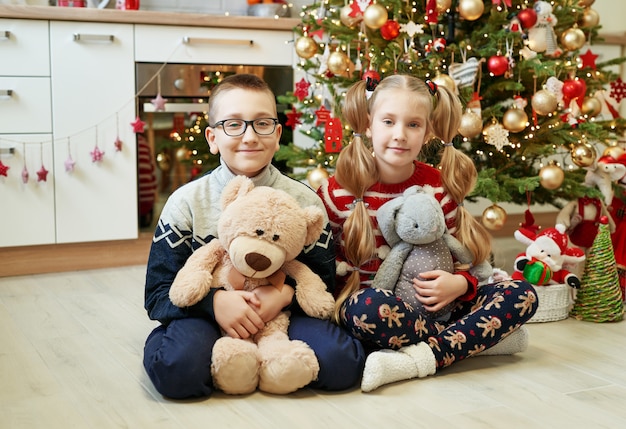 Broer en zus zitten in de buurt van de kerstboom met knuffels