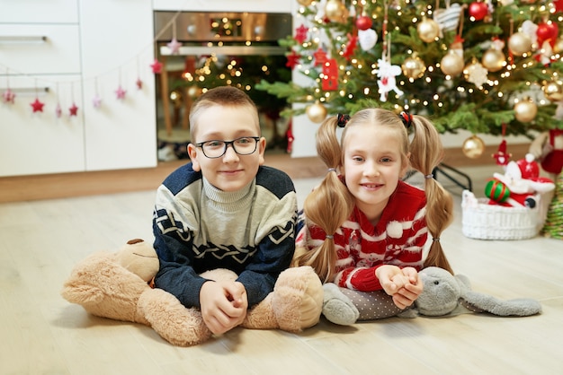 Broer en zus zitten in de buurt van de kerstboom met knuffels