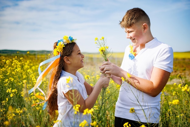 Broer knuffelt zus met Oekraïense krans en boeket bloemen in handen in koolzaadveld onder de hemel