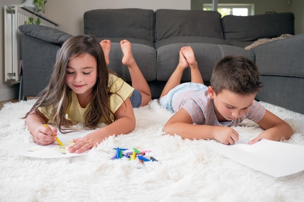 Broers en zussen spelen samen thuis kleine jongen en meisje liggend op het tapijt en tekenen op witte vellen papier met kleurrijke kleurpotloden