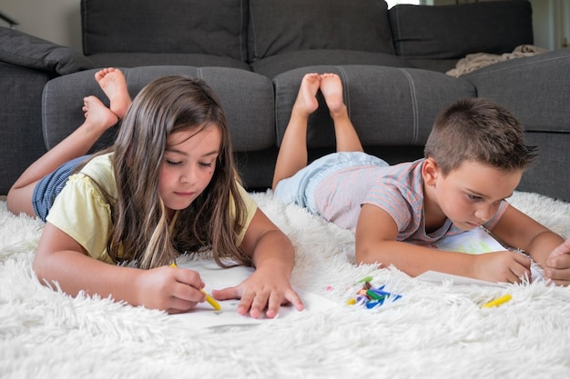 Foto broers en zussen spelen samen thuis kleine jongen en meisje liggend op het tapijt en tekenen op witte vellen papier met kleurrijke kleurpotloden