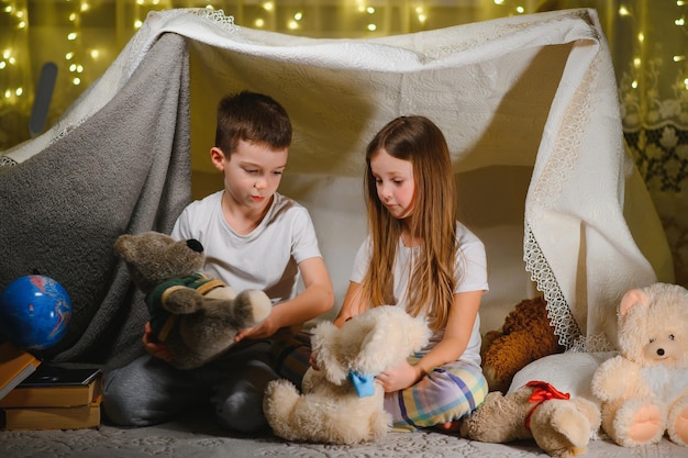 Broers en zussen zitten in een hut van stoelen en dekens Broer en zus lezen thuis boek met zaklamp