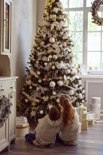 Broertje en zusje omhelzen elkaar en kijken met vreugde en dromen naar een prachtig versierde kerstboom, doen wensen