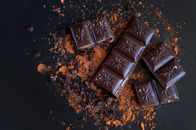 Сломанные кусочки горького шоколада и какао-порошок на темной поверхности, вид сверху