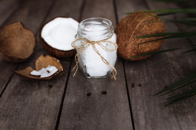 Broken coconuts on gray wooden table with jar of raw organic extra virgin coconut oil and palm leaf. White coconut pulp.