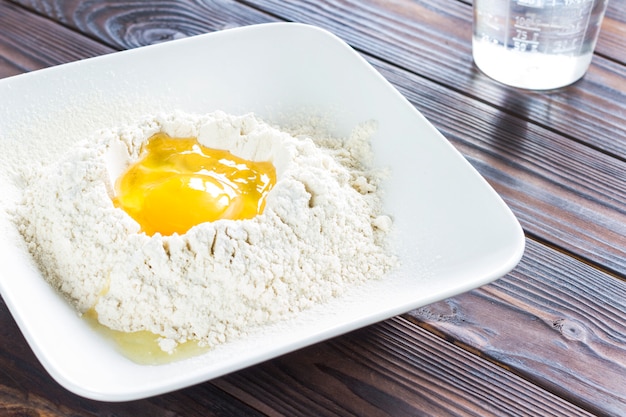 Broken egg in flour in white plate, spoon on plate. Sun's rays on table.