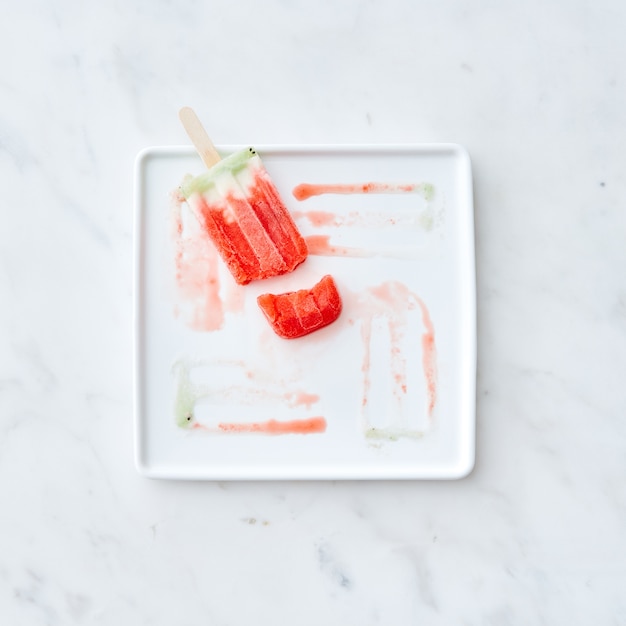 Broken fruit ice cream lolly on a white plate with a pattern from the thawing ice cream on a gray marble background. Copy space for text. Flat lay