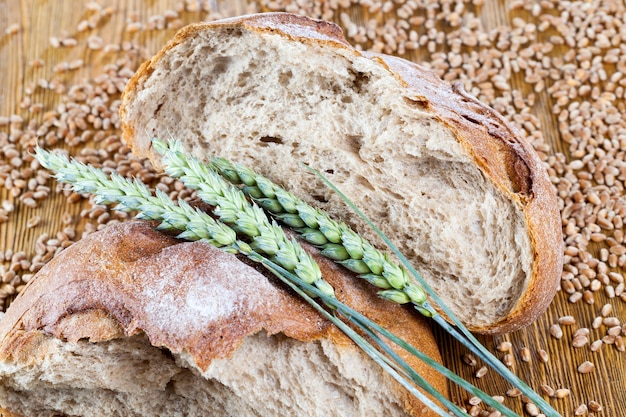 Broken traditional round wheat bread of gray color, with green wheat ears on the board, close-up