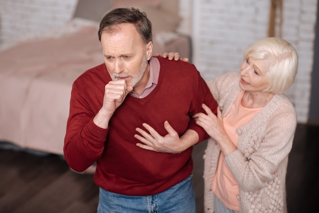 Bronchitis again. Senior man standing and strongly coughing while his wife is giving him support.