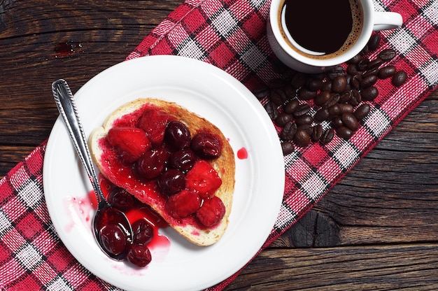 Brood met ingeblikt fruit en koffie Geroosterd brood met ingeblikte kersen, aardbeien en kopje koffie op rustieke houten tafel. Bovenaanzicht