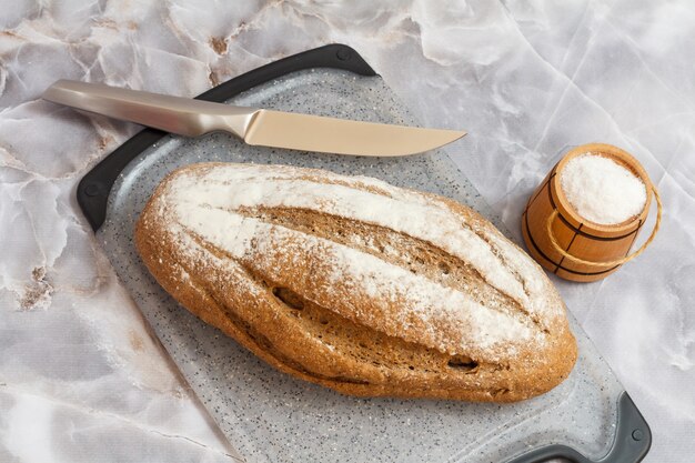 Brood van roggebrood en een keukenmes op een snijplank, een houten zoutkelder met de grijze achtergrond. Bovenaanzicht.