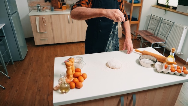 Broodbereiding thuis in de keuken door oude man. Oudere senior bakker met bonete en uniform besprenkelen, zeven, met de hand nieuwe ingrediënten op deeg verspreiden, zelfgemaakte pizza en koekjes bakken