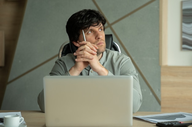 Brooding young man holding pen in hand and looking away while sitting at his desk