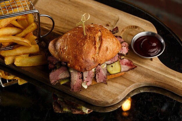 Broodje Pastrami op roggebrood geserveerd met augurk en mosterdsaus Lekkere sandwich met frietaardappelen op donkere achtergrond