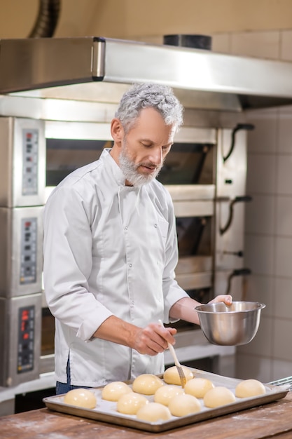 Broodjes koken. Ernstige geïnteresseerde man in wit uniform die deeg bedekt met speciaal glazuur aanraken van oppervlak van broodje met borstel