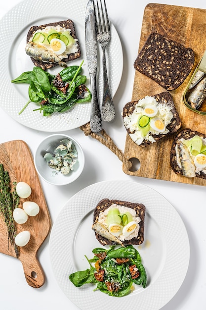 Broodjes met sardientjes, ei, komkommer en roomkaas, salade garnituur met spinazie en gedroogde tomaten. Witte achtergrond. Bovenaanzicht.